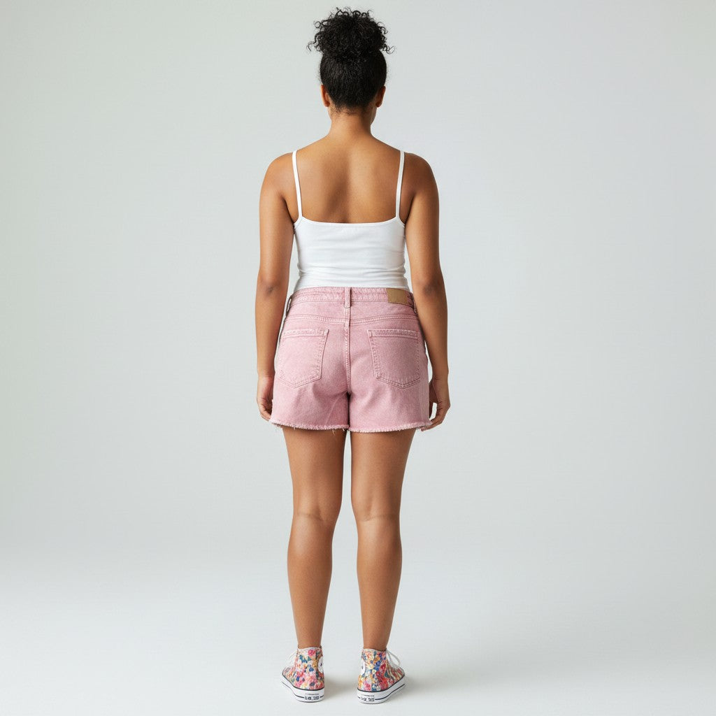 A woman modeling a white spaghetti-strap tank top and light pink denim shorts with distressed details, paired with colorful sneakers, against a light gray studio background.