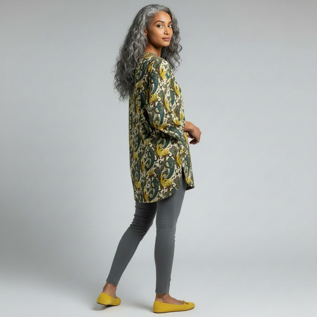 A woman with long gray hair is shown from the side in a studio setting, wearing a loose long-sleeve tunic top with a green, yellow, and cream paisley pattern, gray leggings, and yellow flats against a light gray background.