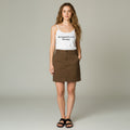 A woman modeling a white spaghetti-strap tank top with 'Reclaimed Revival Boutique' printed in black on the chest, paired with a brown mini skirt and black slide sandals, against a light gray studio background.