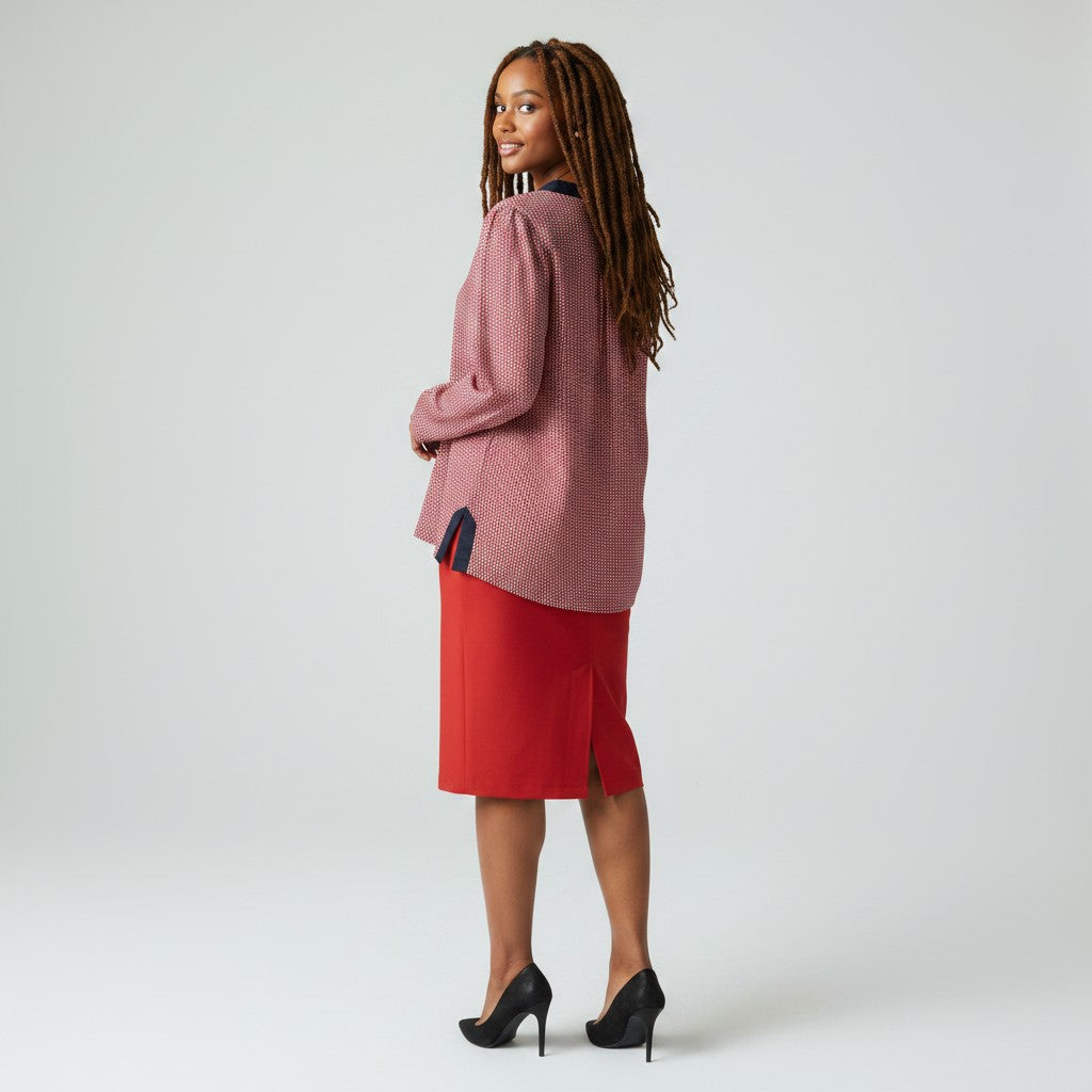 A woman stands in profile wearing a textured dusty rose cardigan with dark navy trim, a bright red midi skirt, and black high heels against a light gray studio background.