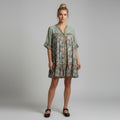 A woman modeling a short-sleeved, tiered bohemian-style dress with a green and beige floral pattern, paired with black Mary Jane shoes, against a neutral gray studio background.