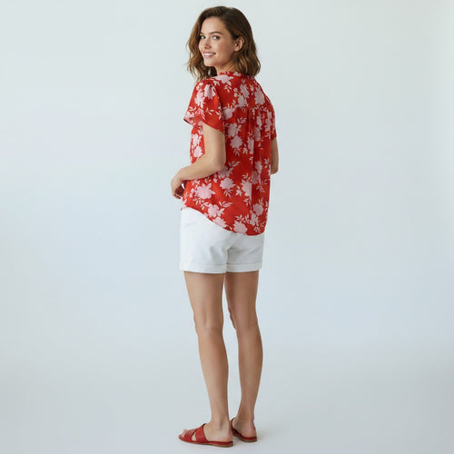 A woman modeling a red short-sleeve top with a white floral pattern, paired with white shorts and red slide sandals, photographed against a light gray studio background.