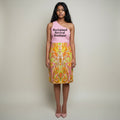 A woman stands against a plain light beige studio background wearing a sleeveless pink top with black text reading 'Reclaimed Revival Boutique' and a knee-length skirt with a bright paisley print in orange, yellow, green, and pink. She is wearing pink pointed heels.