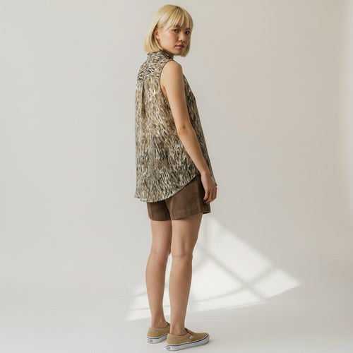 A woman stands in profile wearing a sleeveless, high-low button-up shirt with an abstract animal print in beige, brown, and black, paired with brown shorts and tan slip-on shoes, against a warm off-white studio background.