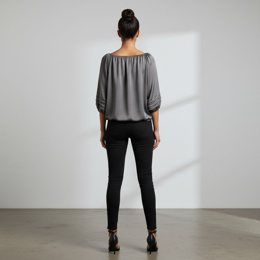 A woman viewed from behind wearing a gray off-the-shoulder blouse with three-quarter sleeves and black skinny pants, standing in a minimalist studio with a white wall and concrete floor.