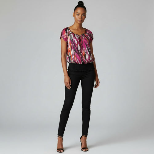 A woman modeling a short-sleeve top with a vibrant pink, orange, and black abstract print, paired with black skinny jeans and black high-heeled sandals, against a light gray studio background.