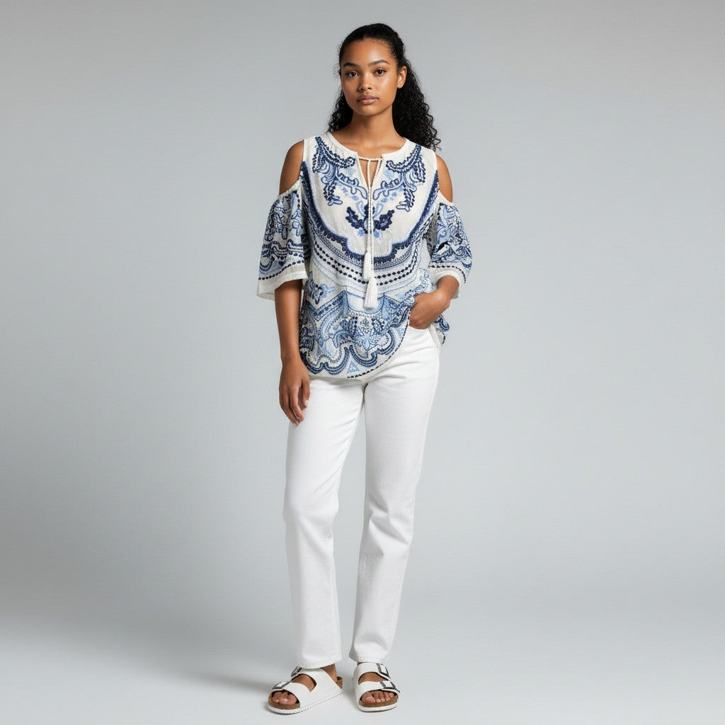 A woman wearing a blue and white floral shirt, white jeans, and white sandals on a studio gray background.