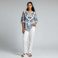 A woman wearing a blue and white floral shirt, white jeans, and white sandals on a studio gray background.