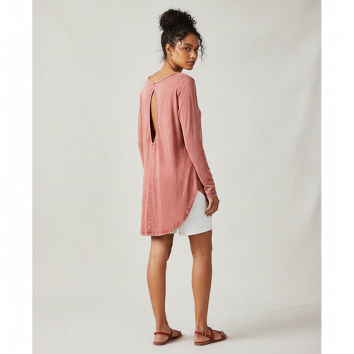 A woman modeling a dusty rose long-sleeve tunic top with a back cutout, paired with white shorts and brown sandals, photographed against a neutral light gray studio backdrop.
