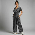 A woman modeling a black and white vertically striped short-sleeve wrap jumpsuit with wide legs, standing against a neutral gray studio background.