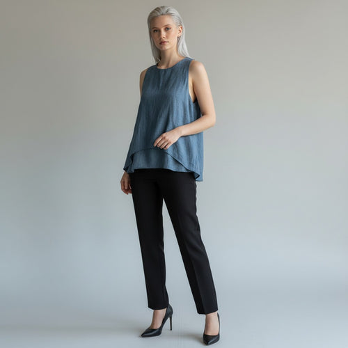 A woman modeling a sleeveless, high-low tiered top in a dusty slate blue color, paired with black trousers and black heels, against a neutral light gray studio background.