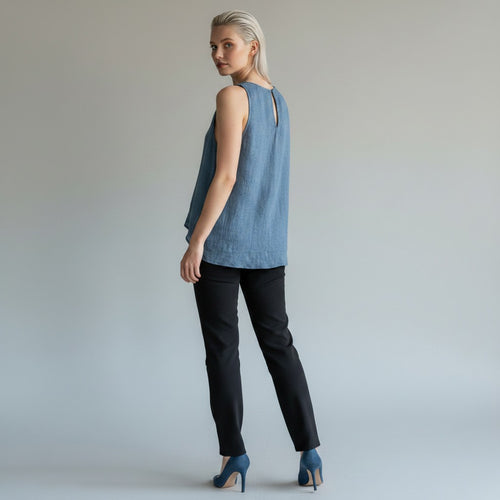 A woman stands in profile wearing a sleeveless light blue top with a small back cutout, black pants, and blue heels against a plain light gray studio background.