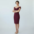 A woman modeling a fitted off-the-shoulder knee-length dress in deep burgundy, paired with nude heels, against a light gray studio background.