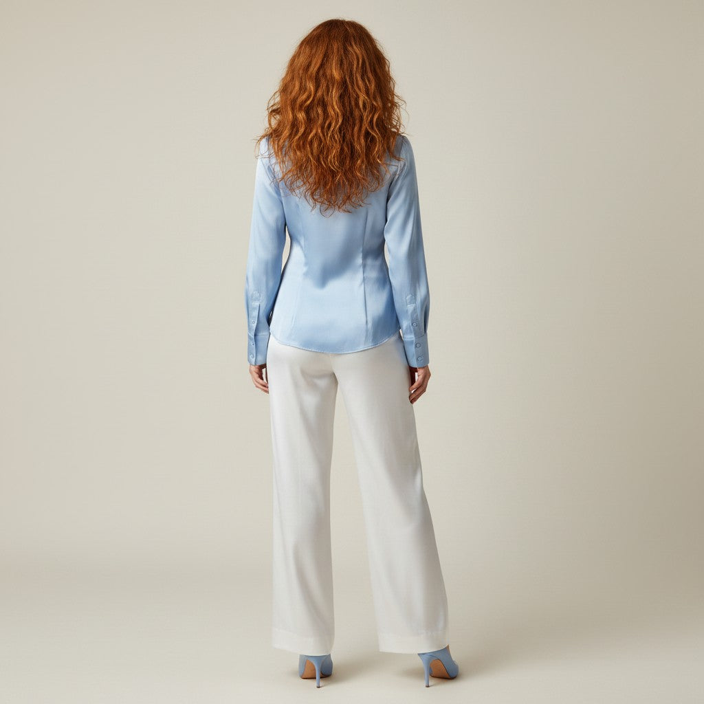 A woman with long wavy red hair is shown from behind wearing a light blue satin long-sleeve blouse and white wide-leg trousers, paired with light blue high heels, against a solid light beige studio background.