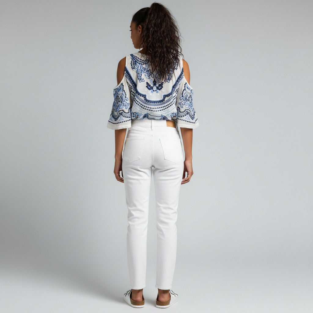 A woman wearing a blue and white floral shirt, white jeans, and white sandals on a studio gray background.
