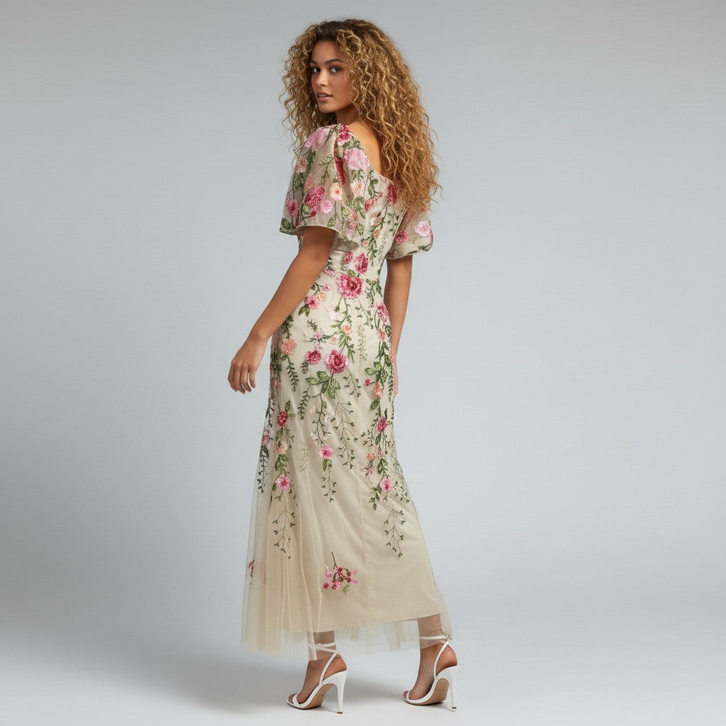 A woman modeling a cream-colored maxi dress with pink floral embroidery and green leaf details, paired with white high-heeled sandals, set against a plain light gray studio background.
