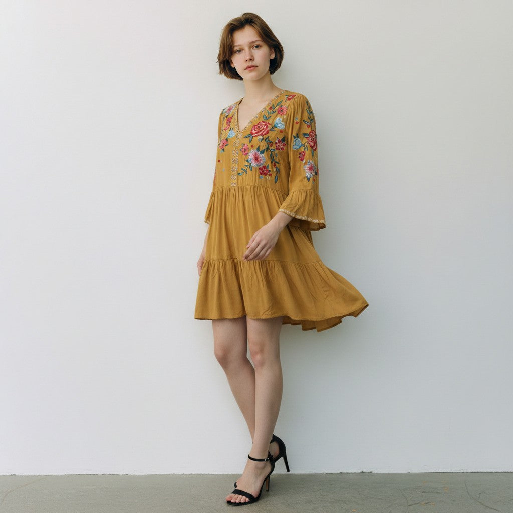 A woman models a tiered mustard yellow dress with colorful floral embroidery on the bodice and sleeves, paired with black strappy high-heeled sandals against a plain white wall.