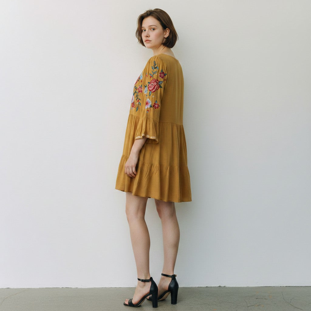 A woman models a tiered mustard yellow dress with colorful floral embroidery on the bodice and sleeves, paired with black strappy high-heeled sandals against a plain white wall.
