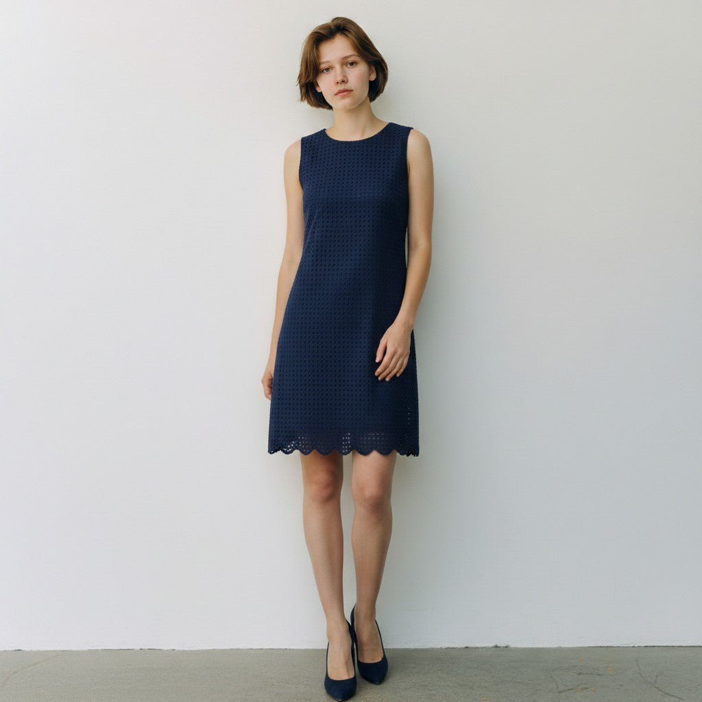 A woman stands against a plain white wall wearing a sleeveless navy blue textured dress with a scalloped hem and dark navy heels.