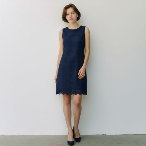 A woman stands against a plain white wall wearing a sleeveless navy blue textured dress with a scalloped hem and dark navy heels.