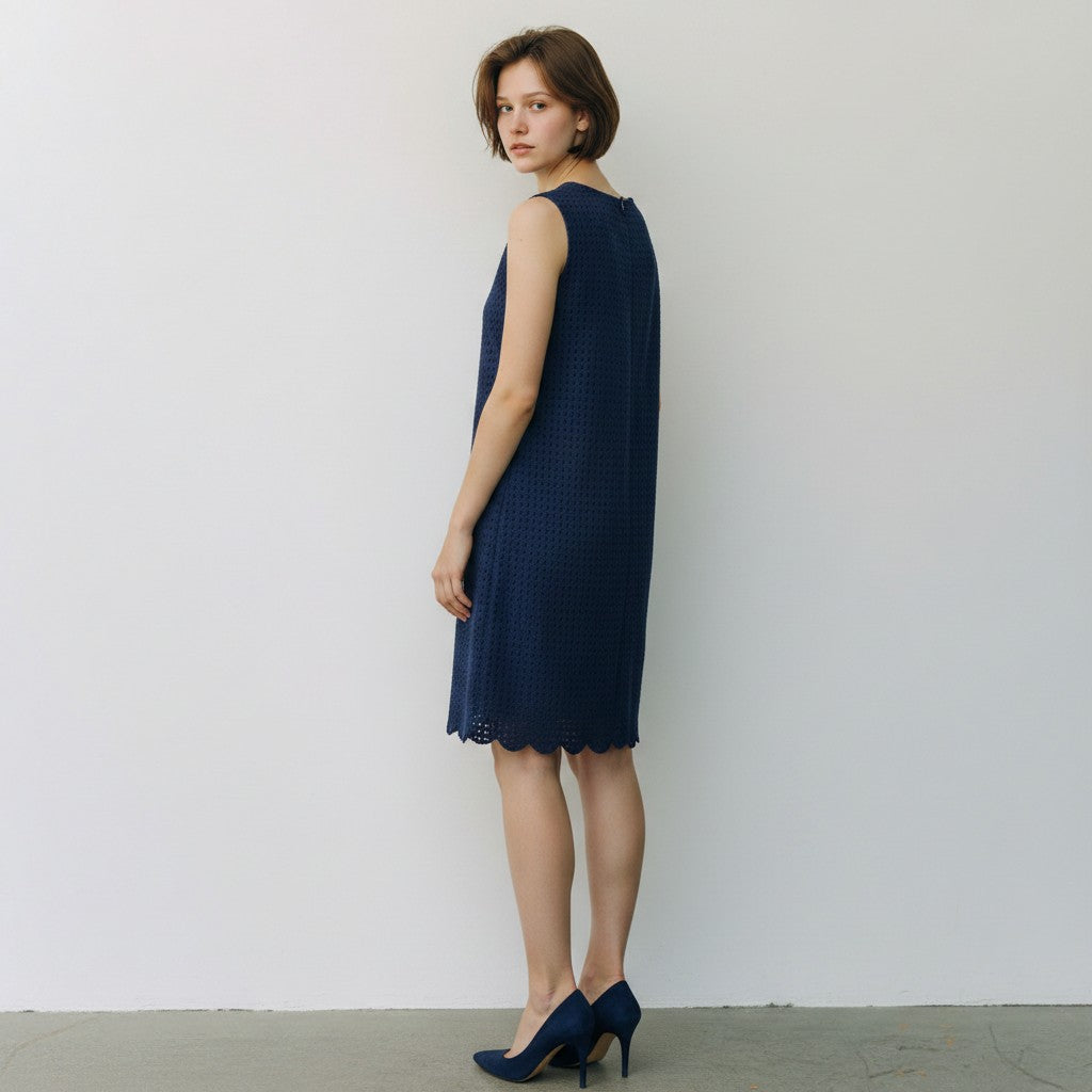 A woman stands in profile wearing a sleeveless navy blue textured knit dress with a scalloped hem and matching navy heels against a plain off-white wall.