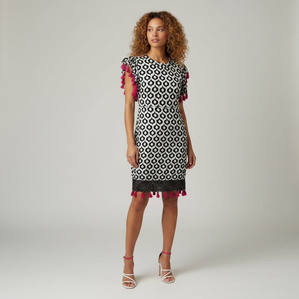A woman modeling a short-sleeve black and white geometric pattern dress with bright pink tassel trim on the sleeves and hem, paired with pink strappy heels, against a light gray studio background.