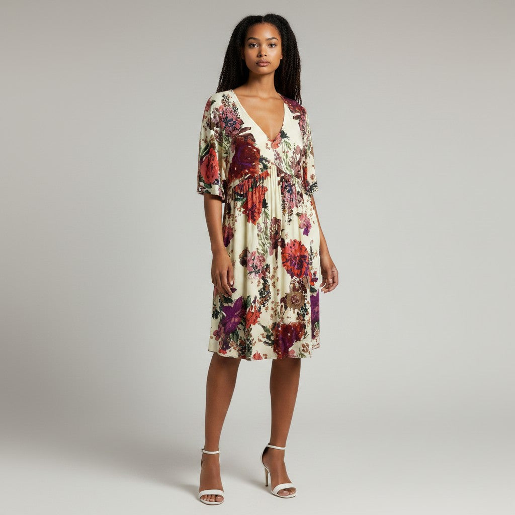 A woman modeling a knee-length wrap-style dress with a cream base and a bold floral print in red, purple, and green, paired with white heeled sandals against a light gray studio background.