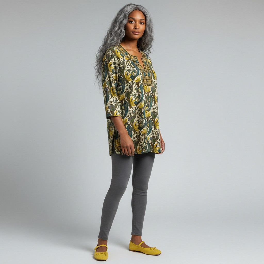 A woman modeling a loose, three-quarter sleeve tunic top with an intricate green, yellow, and beige paisley pattern, paired with gray leggings and yellow flats, against a light gray studio background.
