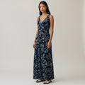 A woman modeling a sleeveless navy maxi dress with a white floral and leaf print, standing against a plain light beige studio background.
