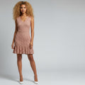 A woman modeling a sleeveless, V-neck lace mini dress in a dusty rose pink color, paired with white pointed-toe heels, against a light gray studio background.