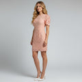 A woman modeling a short peach-pink lace dress with short puff sleeves and white pointed-toe heels, standing against a light gray studio background.