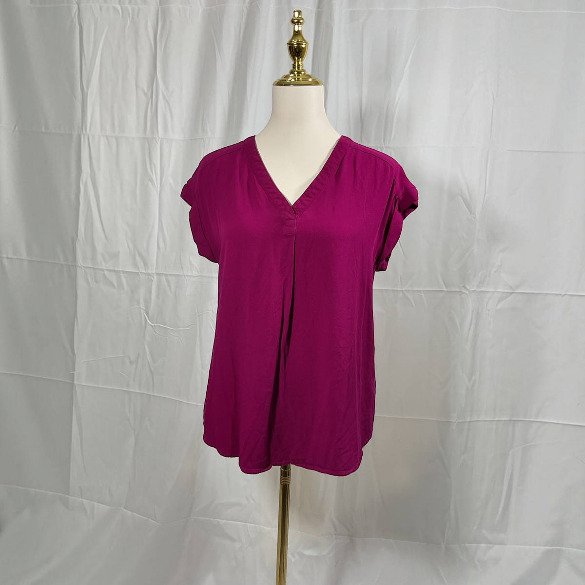 A magenta short-sleeve V-neck blouse displayed on a cream dress form with a gold finial, set against a wrinkled white fabric backdrop.