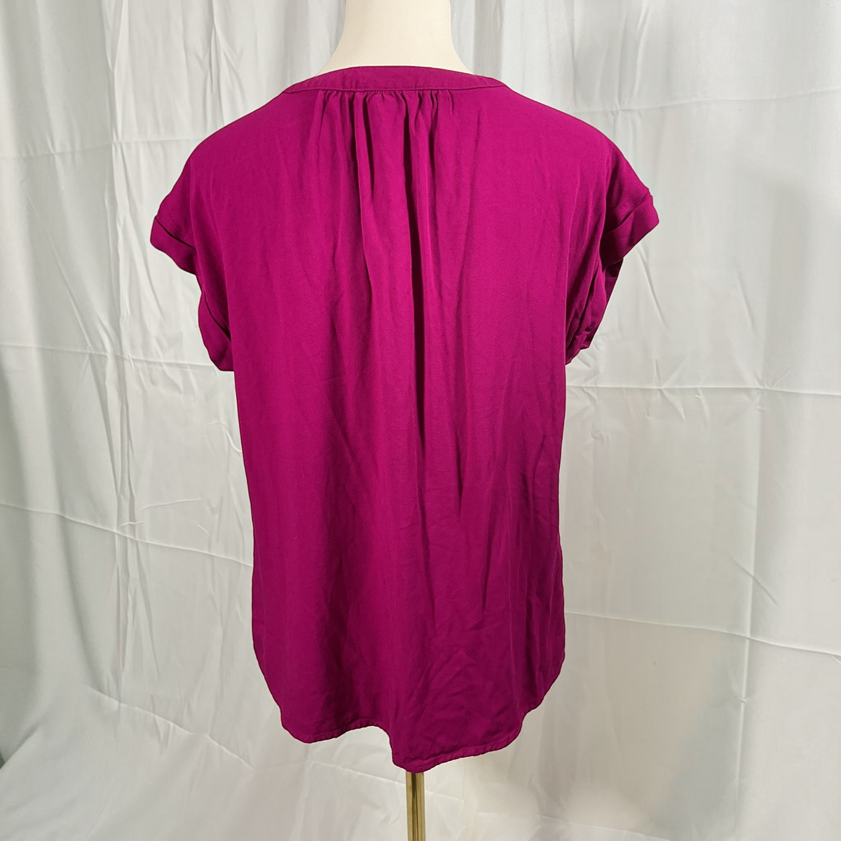 A magenta short-sleeve top displayed on a mannequin torso, showing the back view with a gathered neckline and vertical seam, set against a wrinkled white fabric backdrop.