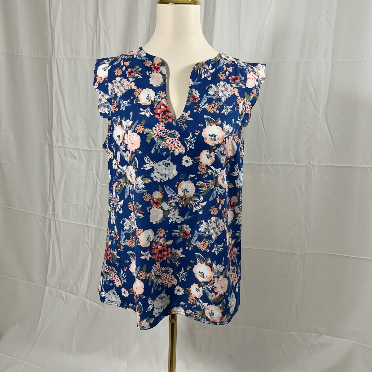 A sleeveless navy blue blouse with a dense floral print in pink, white, and red, displayed on a cream mannequin torso against a wrinkled white fabric backdrop.