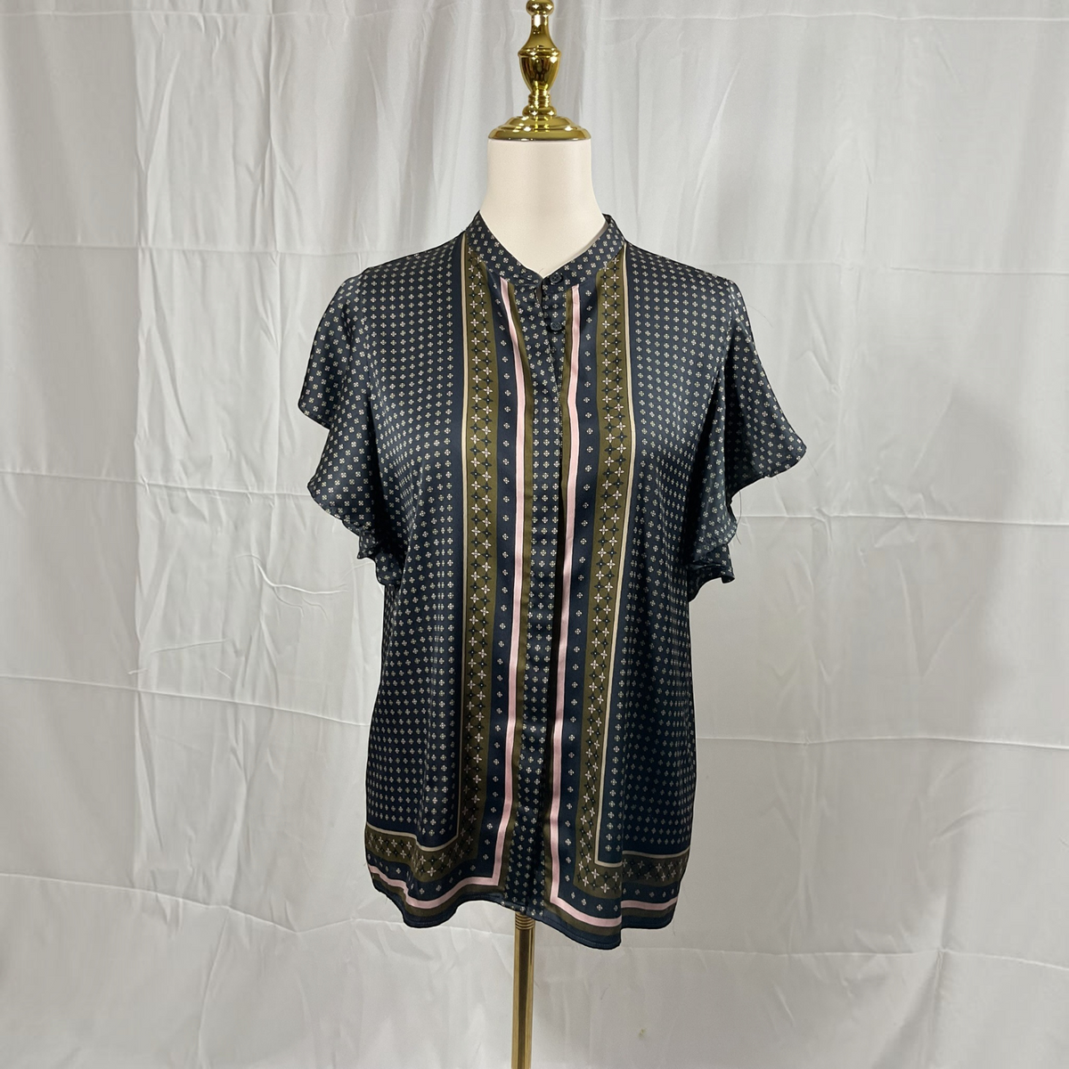 A woman stands in profile wearing a black short-sleeve blouse with a subtle geometric pattern and a decorative gold and beige border, paired with black skinny pants and white high-heeled sandals, against a light gray studio background.