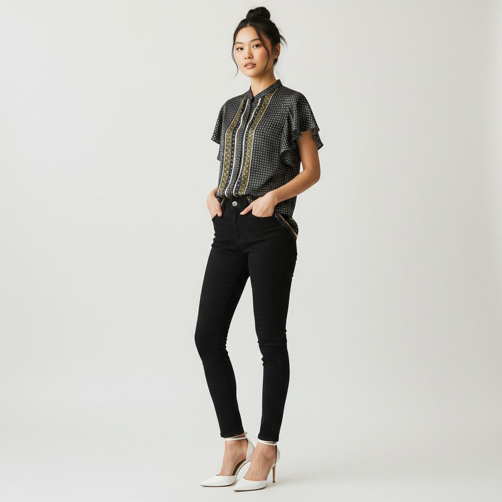 A woman stands in profile wearing a black short-sleeve blouse with a subtle geometric pattern and a decorative gold and beige border, paired with black skinny pants and white high-heeled sandals, against a light gray studio background.