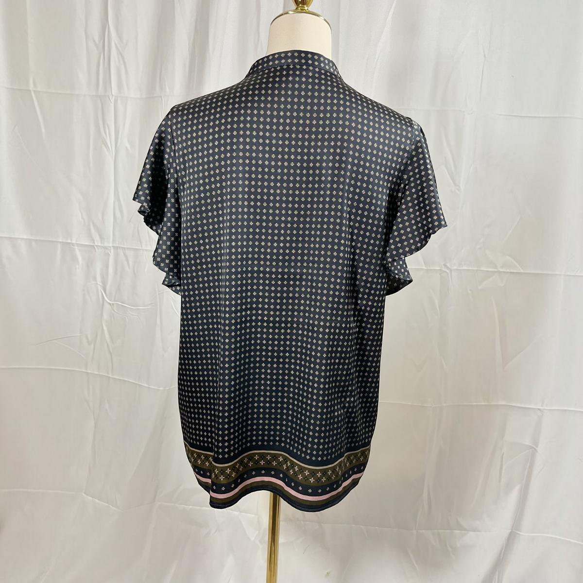 A woman stands in profile wearing a black short-sleeve blouse with a subtle geometric pattern and a decorative gold and beige border, paired with black skinny pants and white high-heeled sandals, against a light gray studio background.