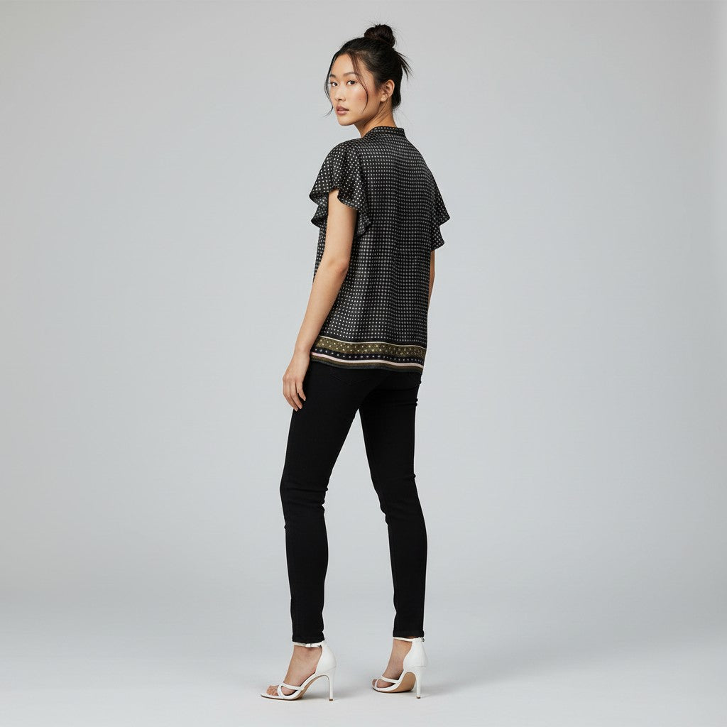 A woman stands in profile wearing a black short-sleeve blouse with a subtle geometric pattern and a decorative gold and beige border, paired with black skinny pants and white high-heeled sandals, against a light gray studio background.