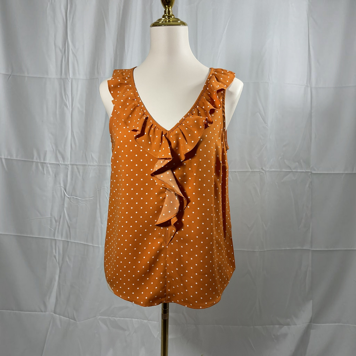 A woman modeling a sleeveless rust-orange top with a small white polka dot pattern and ruffle trim, paired with black skinny pants and white heeled sandals, against a plain light gray studio background.