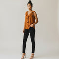A woman modeling a sleeveless rust-orange top with a small white polka dot pattern and ruffle trim, paired with black skinny pants and white heeled sandals, against a plain light gray studio background.