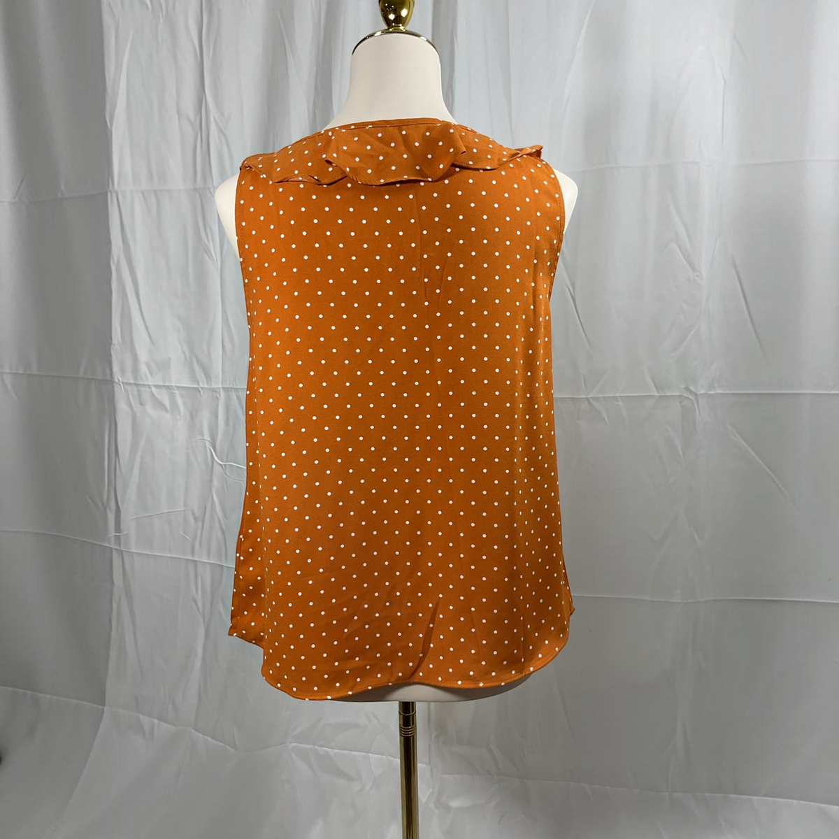 A woman modeling a sleeveless rust-orange top with a small white polka dot pattern and ruffle trim, paired with black skinny pants and white heeled sandals, against a plain light gray studio background.