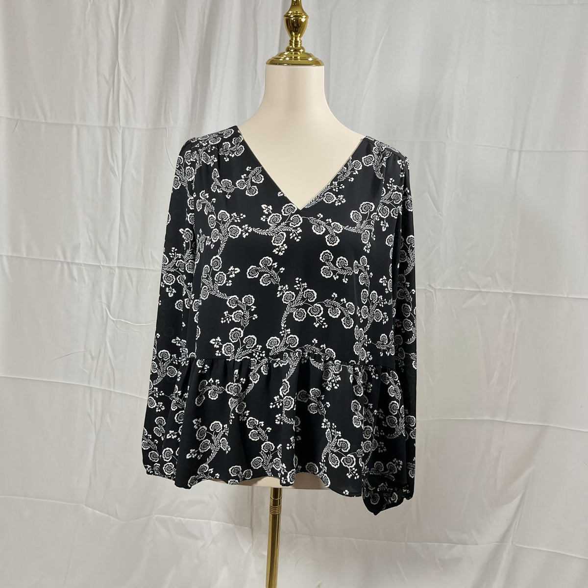 A black long-sleeve blouse with a white floral pattern, displayed on a white mannequin torso against a white fabric backdrop.