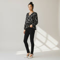 A woman stands in a studio wearing a black long-sleeve blouse with a white floral pattern, black skinny pants, and white high heels. The background is a light neutral studio wall with soft window-like shadows.