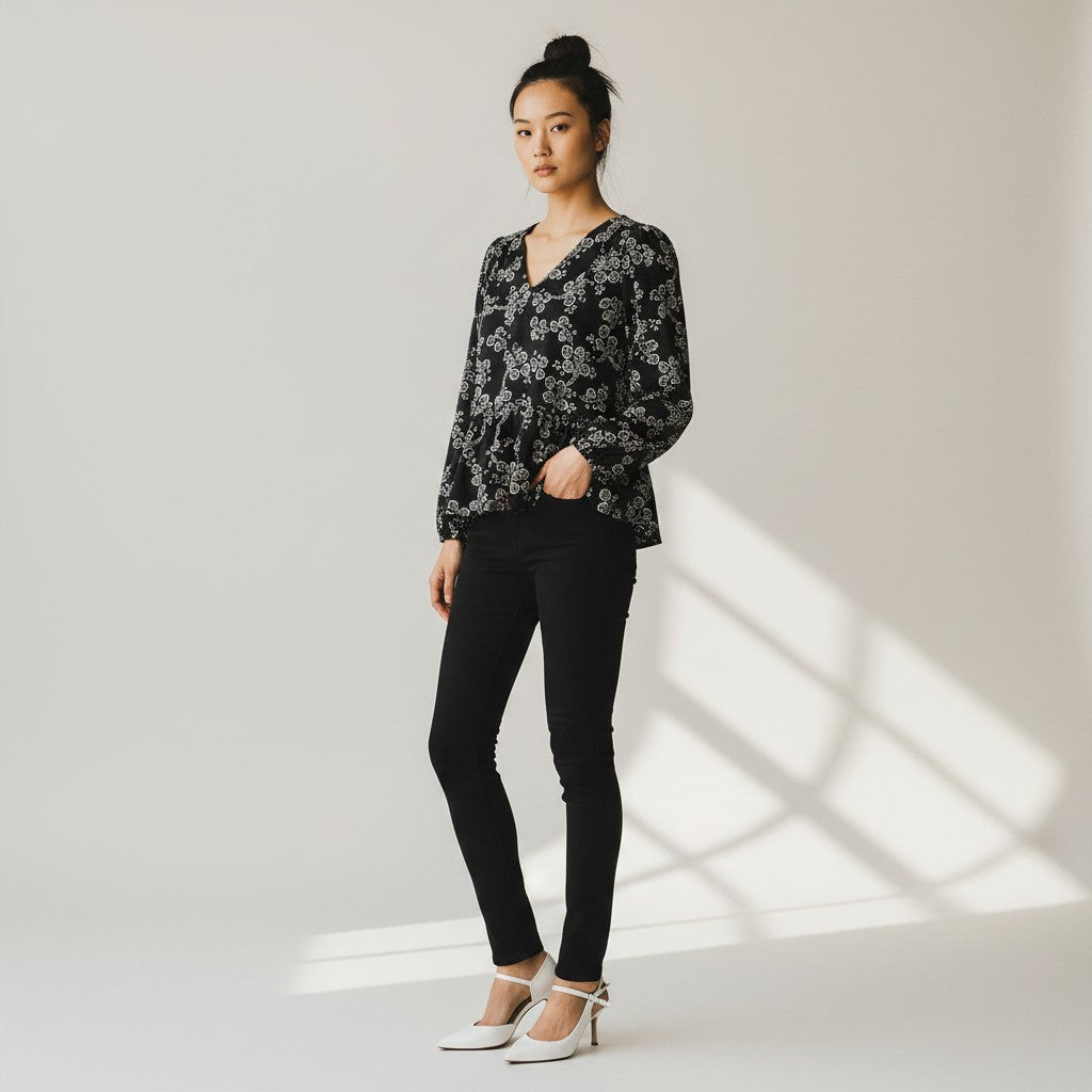 A woman stands in a studio wearing a black long-sleeve blouse with a white floral pattern, black skinny pants, and white high heels. The background is a light neutral studio wall with soft window-like shadows.