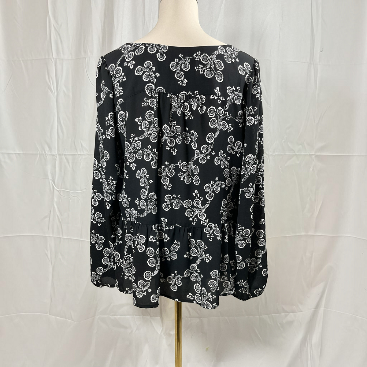 A black long-sleeve blouse with a white floral pattern, displayed on a mannequin torso against a white wrinkled fabric backdrop.