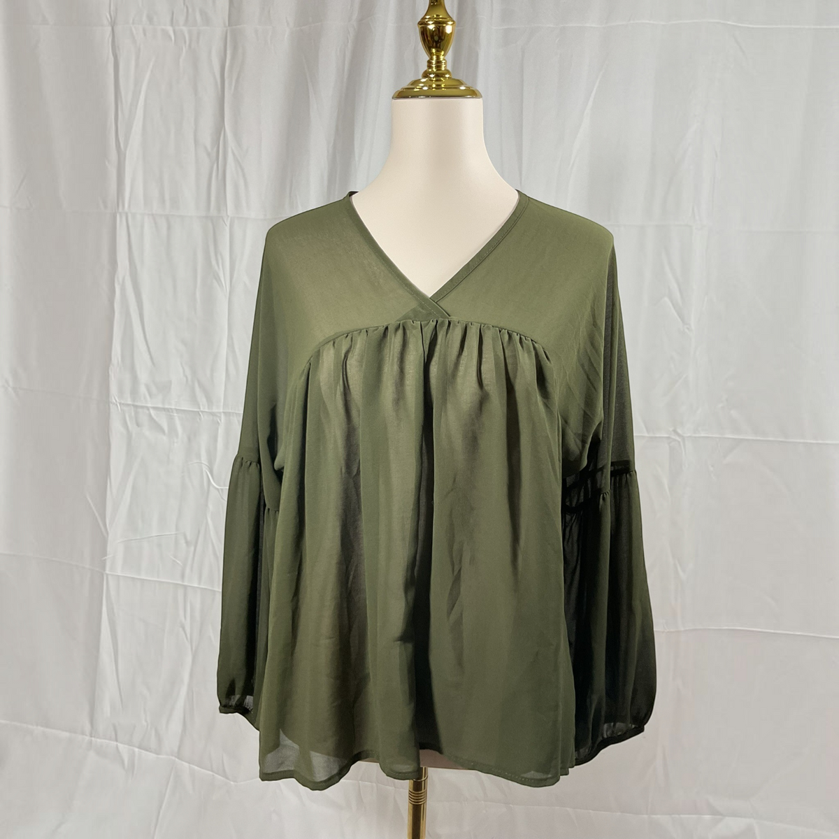 An olive green long-sleeve blouse with a V-neckline and gathered bust detail, displayed on a cream dress form with a gold finial against a white fabric backdrop.