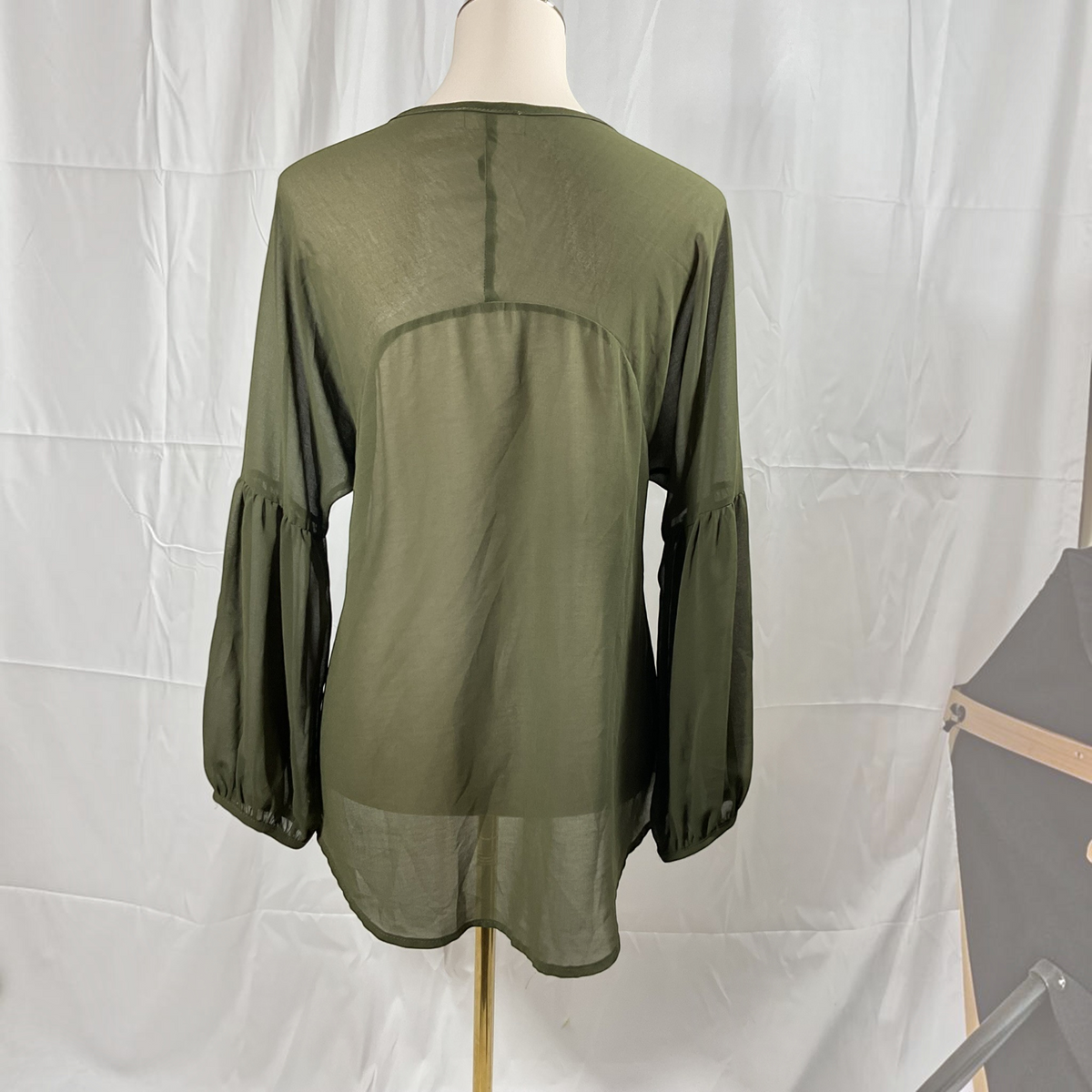 An olive green sheer long-sleeve blouse displayed on a mannequin torso against a white fabric backdrop.