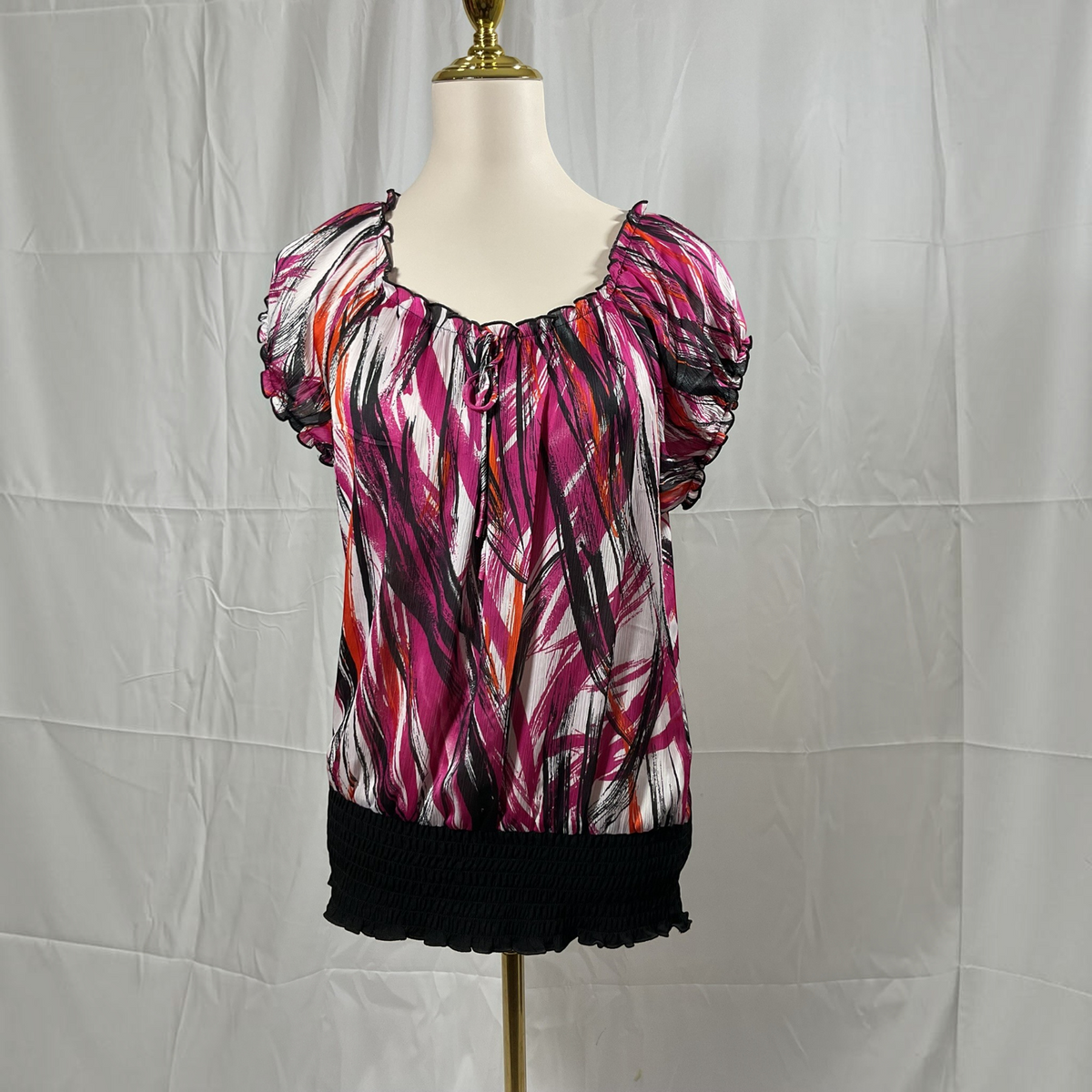 A short-sleeve top with an abstract brushstroke pattern in pink, black, white, and orange, displayed on a cream mannequin torso against a wrinkled white backdrop.