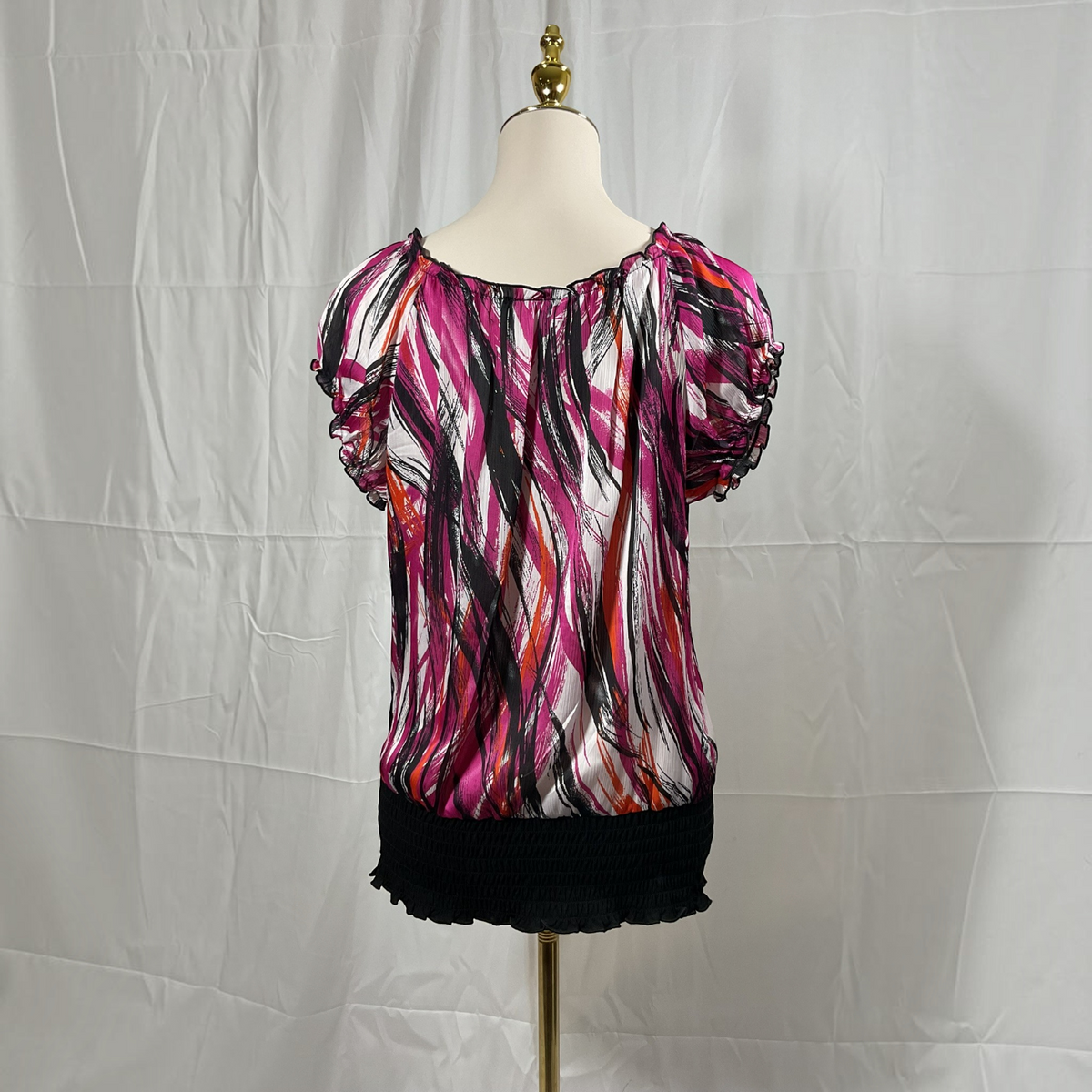 A short-sleeve blouse with puff sleeves and an abstract brushstroke print in pink, black, and orange on a white base, displayed on a dress form against a white fabric backdrop.