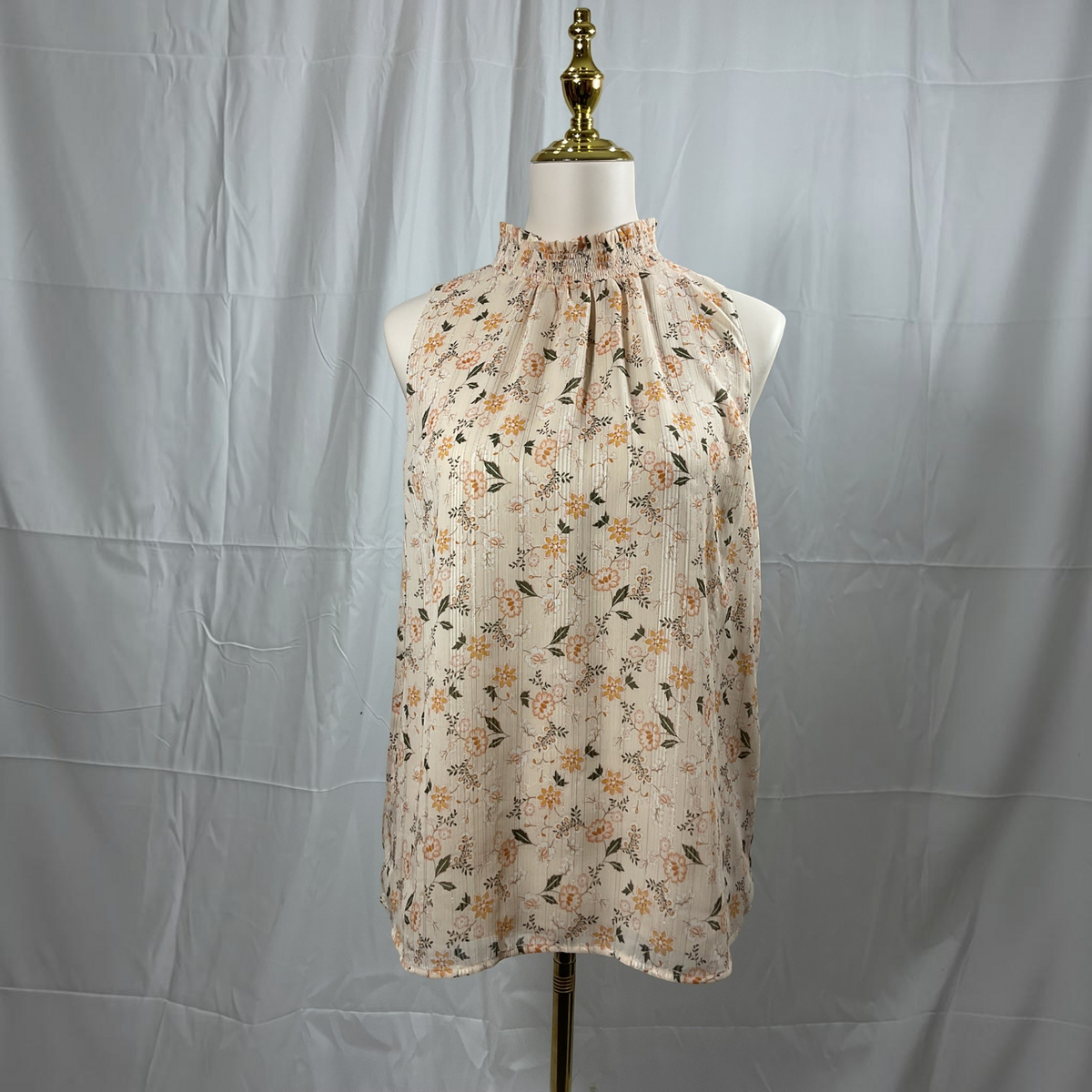 A sleeveless high-neck blouse with a small-scale floral print, displayed on a mannequin torso against a wrinkled light gray fabric backdrop.